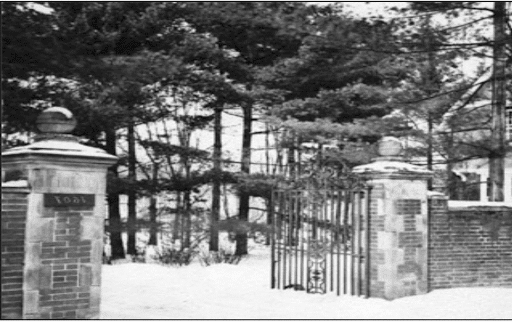 Black and White photo of a wrought iron gate with brick pillars on either side connected to brick walls. Trees fill the space behind the gate, which is open.