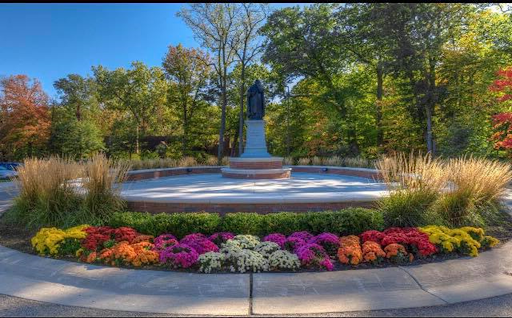 Statue of St. Thomas Aquinas at the center of a paved circle surrounded by a short brick wall lined with bushes, flowers, and grasses. Trees line the backdrop.