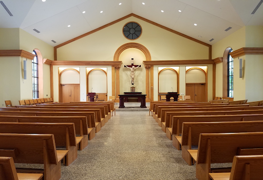 A view down the aisle of Our Lady Seat of Wisdom Chapel. Warm wood pews on either side. Christ on the Cross looms over the alter. The walls are a yellowish beige lined with warm wood details.